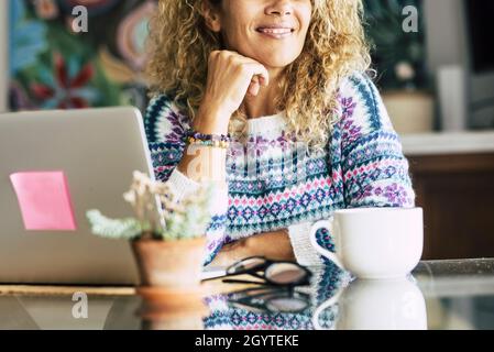 Happy young woman smiling and relaxing with hand on chin while working on laptop on table. Businesswoman working from home. Thoughtful young woman in Stock Photo