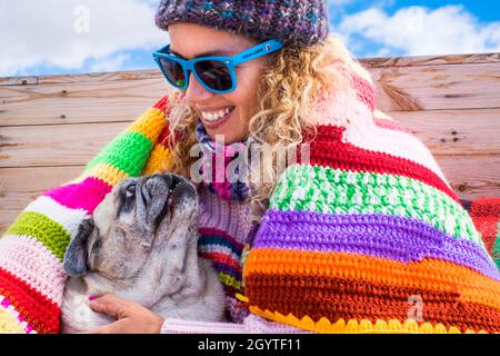 Young woman embracing her pet dog while relaxing on bed against sky. Close up of woman and her dog pug in winter clothes. Happy woman with pet enjoyin Stock Photo