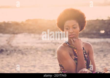 Thoughtful african american woman sitting on beach with hand on chin. Attractive black woman relaxing on sandy beach during holidays. Young woman spen Stock Photo
