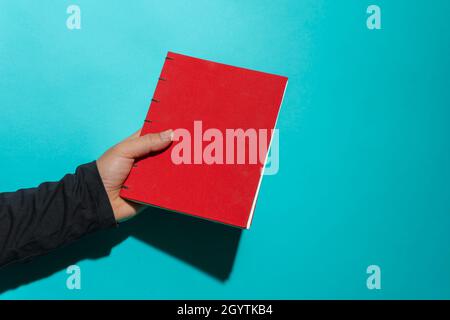 Hand holding red book with clean cover in front of blue background with copy space for text. Stock Photo