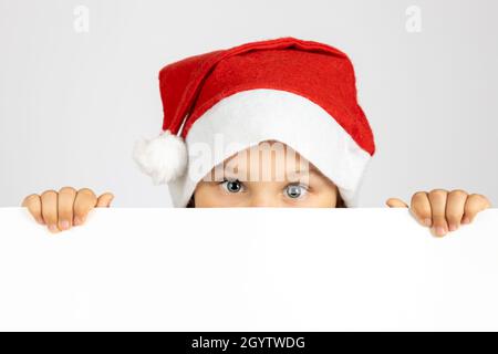 portrait of cute child in red Santa Claus hat hiding face behind white blank banner isolated on white background with copy space Stock Photo