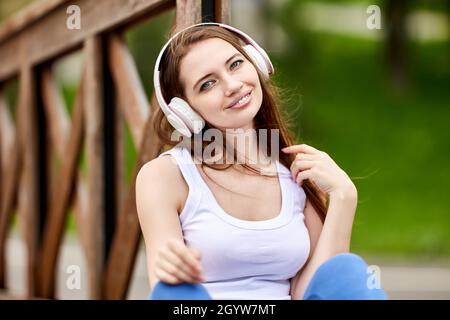 Smiling young woman in wireless headphones sits outdoors. Stock Photo