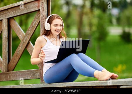 Remote work by happy white woman in headphones with laptop outdoors. Stock Photo