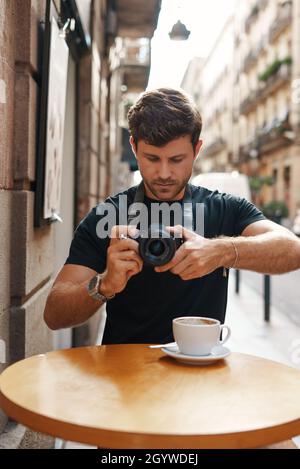 Concentrated young male photographer focusing and taking picture on modern digital camera while sitting at wooden table in street cafe during coffee break Stock Photo