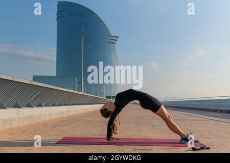 Side view of young woman in black activewear doing Purvottanasana pose against blue sky and skyscraper on city street. Stock Photo