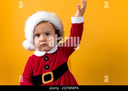 Serious Portraite Cute Happy Cheerful Chubby Baby Girl in Santa Hat Waving Hand Up At Yellow Background. Child Play Christmas Scene Celebrating Stock Photo