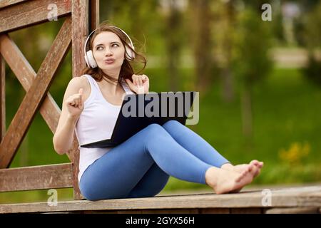 Grimacing woman in cordless headphones with laptop sits on nature. Stock Photo