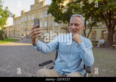 Smiling senior man, recovering male patient in wheelchair making a video call using smartphone while spending time alone in the park near hospital Stock Photo