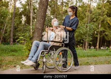 Young nurse in face shield and protective face mask assisting senior handicapped man in wheelchair using his smartphone during a walk in the park Stock Photo