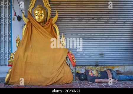 An apparently drunk man sleeps next to a Buddha statue placed outside a shop for Buddhist paraphernalia; Bamrung Muang Road, Bangkok, Thailand Stock Photo