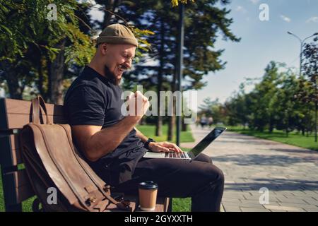 happy victorious bearded man sit on the bench outdoor while look at laptop gesturing yes win winner  Stock Photo