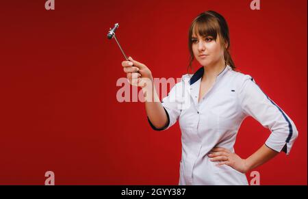 A doctor in a white coat holds a neurological hammer on a red background to test reflexes. Stock Photo