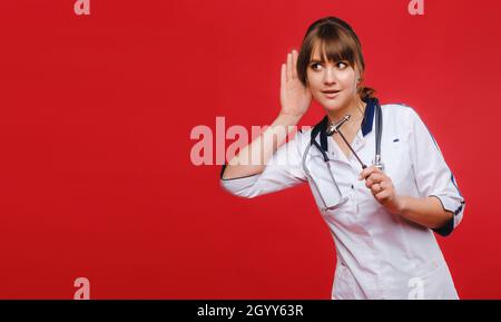A doctor in a white coat on a red background holds a neurological hammer and listens to something. Stock Photo