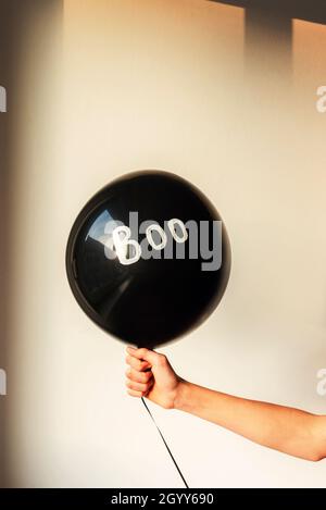 Girl's hand holding black baloon with word Boo on white wall background. Halloween concept. Stock Photo