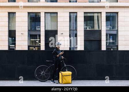 Side view of delivery woman holding a mobile phone, wearing a cycling helmet. Young female standing in the city with bicycle and thermal backpack. Stock Photo