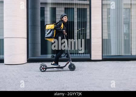Cheerful female courier standing on electric scooter. Woman in cycling helmet wearing thermal backpack looking away. Stock Photo