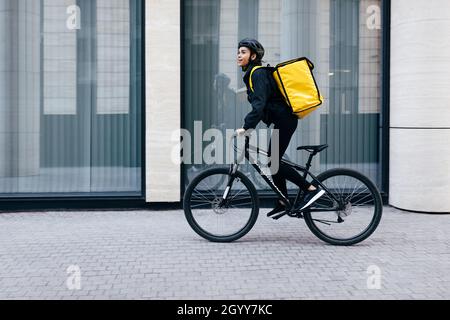 Side view of a young woman riding her bike in the city. Courier with a thermal backpack in her way to deliver food to a customer. Stock Photo