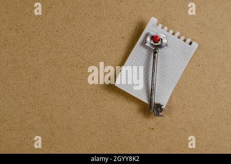 Large metal door key and a piece of paper against a brown background. The old key hangs vertically on a red pin. Torn from a notepad on spring blank p Stock Photo