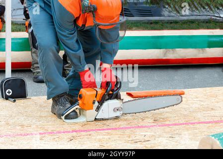 A man starts and tunes a chainsaw against the background of a field of competition between lumberjacks and a marking log. Copy space. Stock Photo