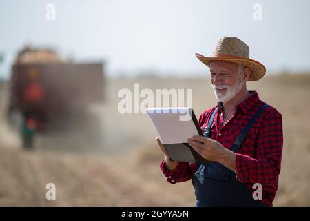 Portrait of senior farmer with straw hat looking at notes in front of tractor with trailer working in corn field during harvest time Stock Photo