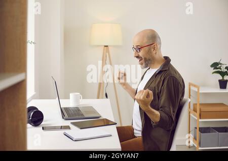 Joyful and happy adult man emotionally rejoices sitting in front of laptop in his home office. Stock Photo