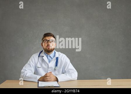 Dreamy young male doctor with a pensive expression looks up sitting at a table on a gray background. Stock Photo