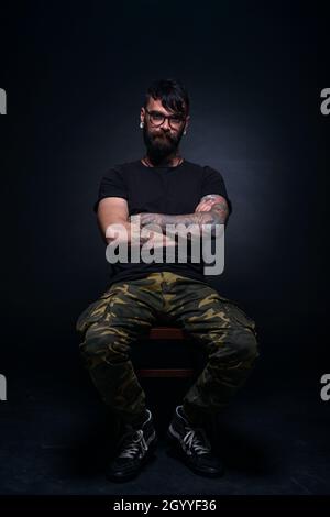Brutal attractive bearded man with tattooed hands poses in black blank t-shirt and army pants seated on a chair Stock Photo