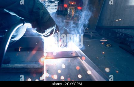 Welder welding metal with argon arc welding machine and has welding sparks and smoke. Man wear protective gloves. Safety in industrial workplace. Stock Photo