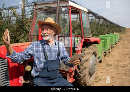 Satisfied senior farmer with tablet standing beside tractor with plastic crates on trailer in modern apple orchard Stock Photo