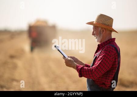 Portrait of senior farmer with straw hat looking at notes in front of tractor with trailer working in corn field during harvest time Stock Photo