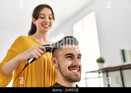 Young hispanic hairdresser woman cutting man's hair using electric razor machine at beauty center. Stock Photo