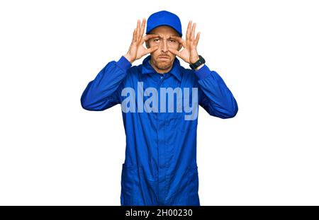 Bald man with beard wearing builder jumpsuit uniform trying to open eyes with fingers, sleepy and tired for morning fatigue Stock Photo