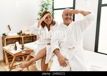 Middle age hispanic couple wearing bathrobe at wellness spa very happy and smiling looking far away with hand over head. searching concept. Stock Photo