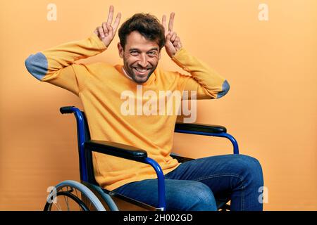 Handsome man with beard sitting on wheelchair posing funny and crazy with fingers on head as bunny ears, smiling cheerful Stock Photo