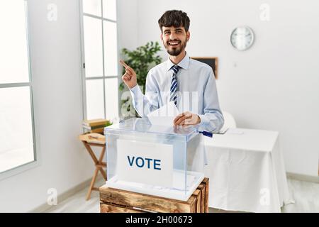 Hispanic man with beard voting putting envelop in ballot box with a big smile on face, pointing with hand finger to the side looking at the camera. Stock Photo
