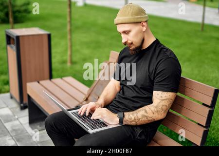 Serious bearded man sits outdoors while typing text working on laptop. 4G internet connection Stock Photo