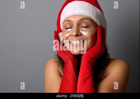 Beautiful woman in Santa carnival outfit, with shiny smoothing spots under her eyes, holds her hands near her face, smiles with a beautiful toothy smi Stock Photo
