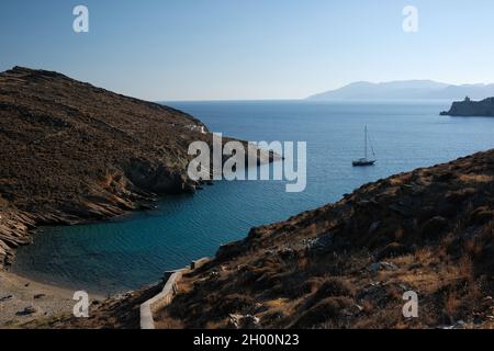 View of the beach of Valmas, a sailboat in the distance and a lighthouse in the backround at the island of Ios in Greece Stock Photo