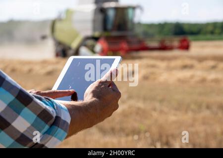 Close up of farmer's hand holding tablet in front of combine harvester working in wheat field Stock Photo