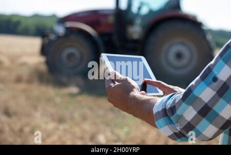 Close up of farmer's hand holding tablet in front of tractor working in wheat field Stock Photo