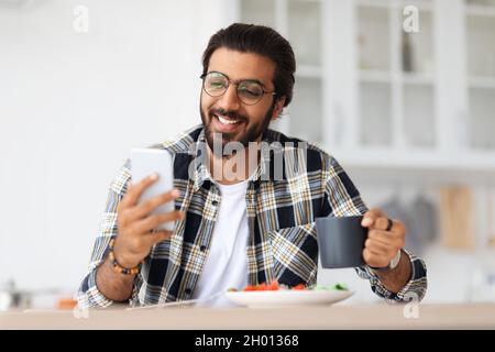 Smiling indian young man eating healthy food, drinking coffee and using modern mobile phone at kitchen, copy space. Happy arab guy having breakfast an Stock Photo