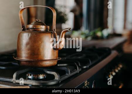 Bronze kettle in modern kitchen. Old vintage teapot on gas stove. Preparing tea. Aluminium teakettle. Sunny daylight from window. Stock Photo