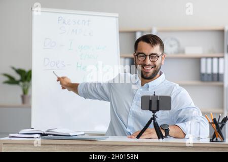 Cheerful adult european male teacher with beard in glasses shows to blackboard and have video call with online class at home interior. Tutor, modern d Stock Photo