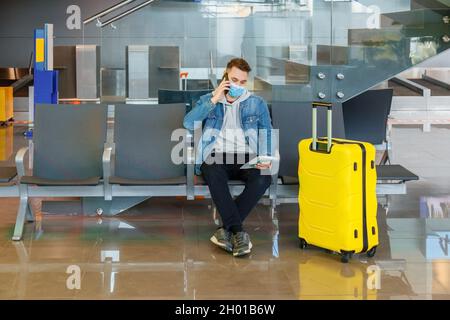 Attractive young man talking on phone in airport lounge with big yellow suitcase before traveling by plane. Caucasian man in Protective face mask is Stock Photo