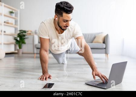 Handsome young Arab guy doing plank, wearing headphones, listening to music, following online video tutorial on laptop at home. Happy Eastern man exer Stock Photo