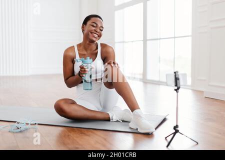 Excited athletic black female in sportswear making video conference with personal trainer or followers, sitting on the floor on yoga mat in front of s Stock Photo