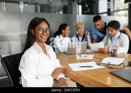Pretty african american young lady doctor smiling at camera while attending breefing, international team of doctors sitting at table at clinic, lookin Stock Photo