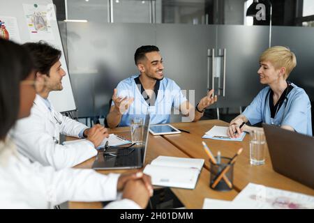 Cheerful doctors men and women multiracial professional medical team having morning breefing, sitting at table, using laptops and having conversation, Stock Photo