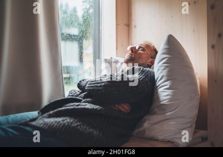 Sleeping Middle-aged man in eyeglasses dressed open cardigan lying on cozy bed pillow next to window. Mental health and autumn weather concept image. Stock Photo