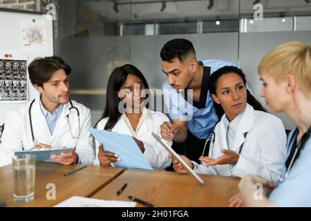 Concentrated multiethnic team of doctors men and women in workwear sitting at table at clinic, having breefing, looking for solutions, discussing clin Stock Photo